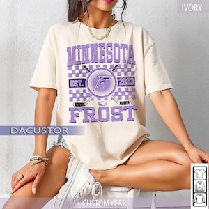 May include: Ivory-colored t-shirt with a purple graphic that says "MINNESOTA FROST" with hockey-themed imagery. The design includes the text "EST. 2023" and a logo. The shirt has short sleeves and a crew neck.