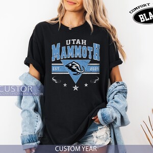 K&ouml;nnte beinhalten: Schwarzes T-Shirt mit dem Text "UTAH MAMMOTH" in Blau und Wei&szlig;, mit einer Mammut-Grafik. Das Shirt enth&auml;lt den Text "EST. 2025" und die Worte "COMFORT COLOR BLACK". Auf der Unterseite des Shirts steht "CUSTOM YEAR".