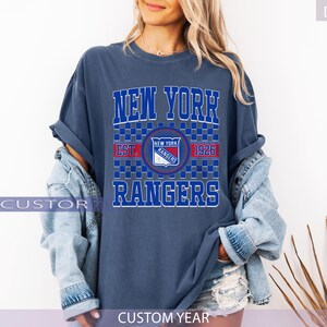 May include: A denim blue t-shirt featuring the text "NEW YORK RANGERS" in white and blue block letters. The shirt includes a team logo and the text "EST. 1926". The shirt is part of a collection of apparel.