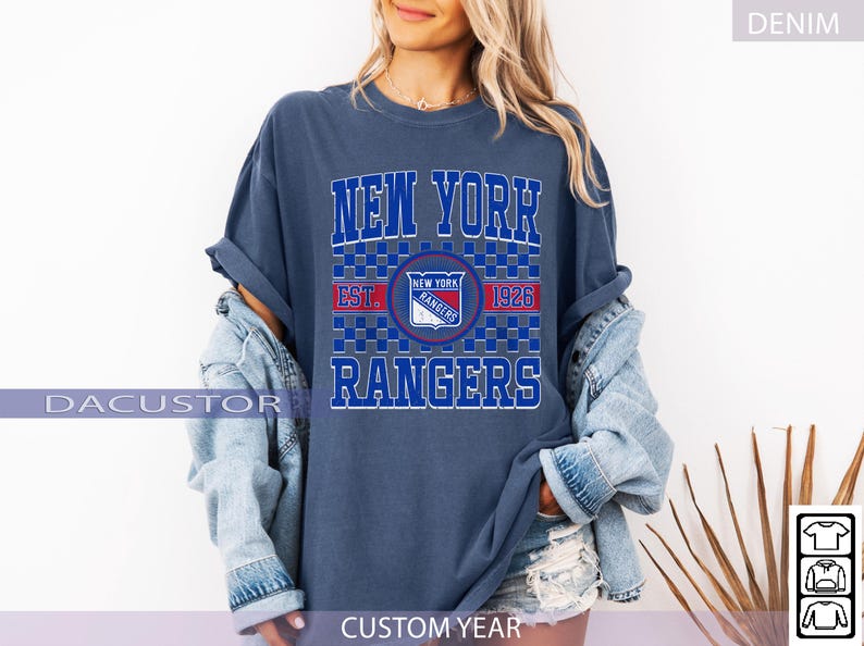 May include: A denim blue t-shirt featuring the text "NEW YORK RANGERS" in white and blue block letters. The shirt includes a team logo and the text "EST. 1926". The shirt is part of a collection of apparel.