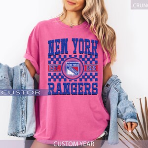 May include: A pink t-shirt with a blue and white graphic that says "NEW YORK RANGERS" and "EST. 1926". The shirt has a crew neck and short sleeves. The model is wearing a denim jacket over the shirt.