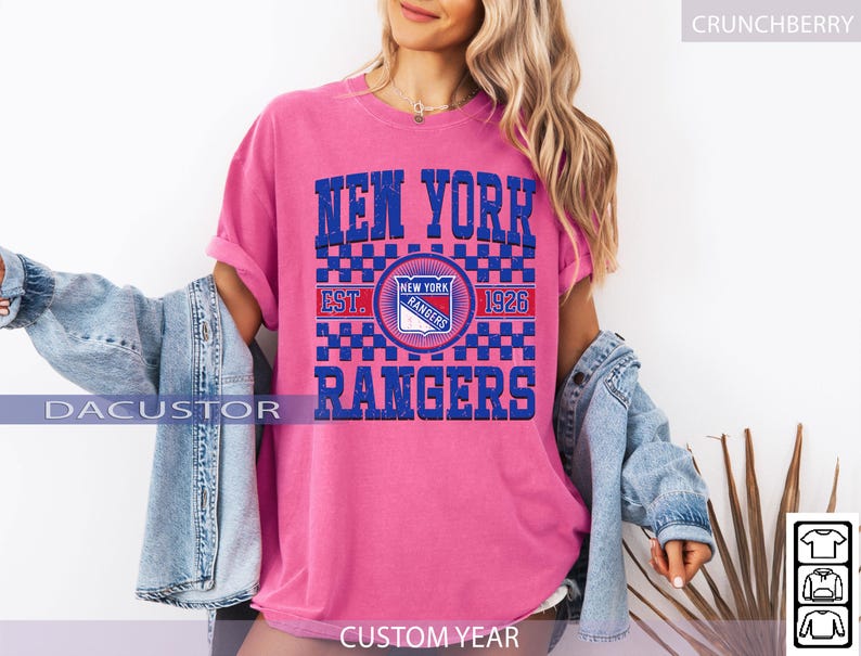 May include: A pink t-shirt with a blue and white graphic that says "NEW YORK RANGERS" and "EST. 1926". The shirt has a crew neck and short sleeves. The model is wearing a denim jacket over the shirt.