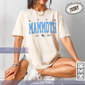 May include: Cream-coloured t-shirt featuring the words "UTAH MAMMOTH" in blue block letters. Below the text is "EST. 2024" and a graphic. The shirt has short sleeves and a crew neck. The words "COMFORT COLOR" and "IVORY" are in a circle.