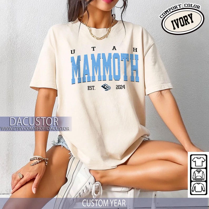 May include: Cream-coloured t-shirt featuring the words "UTAH MAMMOTH" in blue block letters. Below the text is "EST. 2024" and a graphic. The shirt has short sleeves and a crew neck. The words "COMFORT COLOR" and "IVORY" are in a circle.