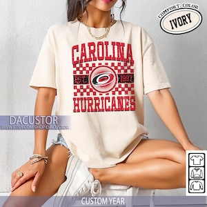 May include: Ivory-colored t-shirt featuring the text "CAROLINA HURRICANES" in red, with a logo and the years "EST. 1997." The shirt has a crew neck and short sleeves. The text "COMFORT COLOR" and "IVORY" are also visible.