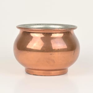 Puede incluir: Una pequeña olla redonda de cobre con un borde plateado. La olla tiene un cuerpo bulboso y una base plana. El cobre tiene una superficie pulida y reflectante. La olla probablemente se utiliza con fines decorativos o para cocinar a pequeña escala.