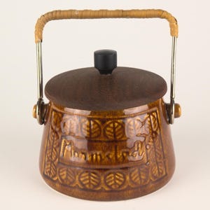 Puede incluir: Tarro de cerámica marrón con tapa y asa trenzada. El tarro presenta un patrón de hojas y la palabra "Montclair" es visible en el lateral. La tapa es de color marrón oscuro con un pomo negro. El asa es de metal y material trenzado.