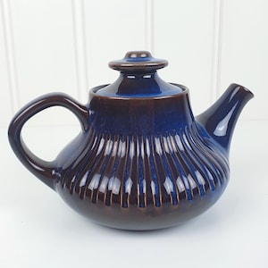 May include: A ceramic teapot with a dark blue and brown glaze. The teapot features a rounded body with vertical ribbed detailing, a curved handle, and a spout. The lid is a matching dark blue colour.