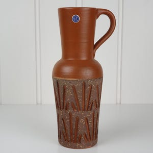 Może przedstawiać: Wysoki dzbanek ceramiczny w kolorze terakoty z gładką górną częścią i teksturowaną podstawą z nakrapianym wykończeniem oraz pionowymi rzeźbionymi liniami. Widoczna jest mała okrągła niebiesko-biała naklejka z napisem Upsala Ekeby Sweden.