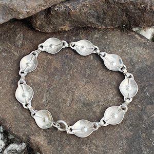 May include: A silver-toned bracelet with a series of oval-shaped links. Each link has a raised, textured design and is connected by smaller, rectangular links. The bracelet is secured with a circular clasp. The jewelry is displayed on a dark, textured surface.