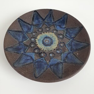 Op de afbeelding: Een rond keramieken bord met een donkerbruine, getextureerde basis. Het midden heeft een lichtblauw en crèmekleurig cirkelvormig patroon omringd door een ring van kleine stippen. Een groot, donkerblauw stervormig ontwerp straalt naar de rand.