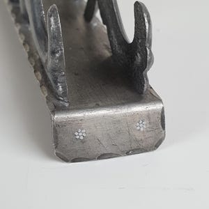 May include: A dark gray metal napkin holder with a hammered texture along the edges. The base features two small, inlaid white flower designs on the front face. The structure has curved, upright supports on each side to hold napkins in place.