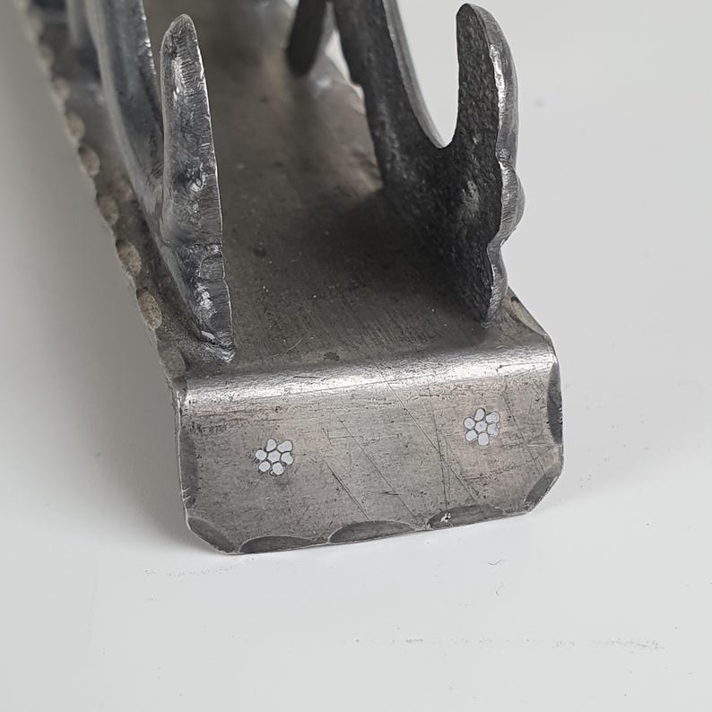 May include: A dark gray metal napkin holder with a hammered texture along the edges. The base features two small, inlaid white flower designs on the front face. The structure has curved, upright supports on each side to hold napkins in place.