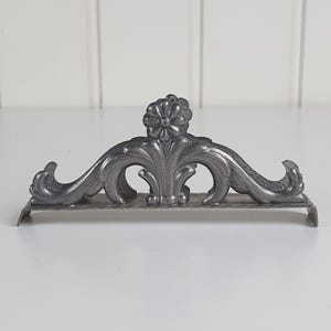 May include: A dark gray metal napkin holder featuring an ornate, symmetrical design with a central flower motif and curved, scrolling acanthus leaf patterns. The structure has a flat base and two raised sides that curve outward at the ends.