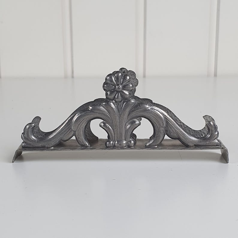 May include: A dark gray metal napkin holder featuring an ornate, symmetrical design with a central flower motif and curved, scrolling acanthus leaf patterns. The structure has a flat base and two raised sides that curve outward at the ends.