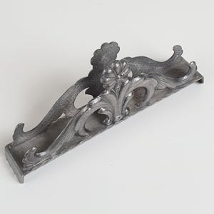 May include: A silver-toned metal napkin holder featuring an elongated, arched design with intricate embossed floral and scrollwork patterns. The structure includes a central flower motif and curved side supports, finished with a textured surface.
