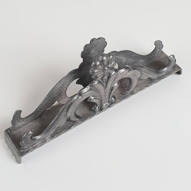 May include: A silver-toned metal napkin holder featuring an elongated, arched design with intricate embossed floral and scrollwork patterns. The structure includes a central flower motif and curved side supports, finished with a textured surface.