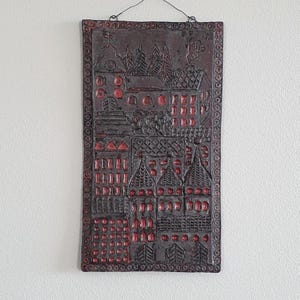 Op de afbeelding: Een rechthoekige donkerbruine keramische wandplaat met een reliëfontwerp van een dorpsgezicht met gebouwen, bomen en een kerk. De architecturale details en ramen zijn geaccentueerd met rood en de rand heeft een cirkelvormig patroon.