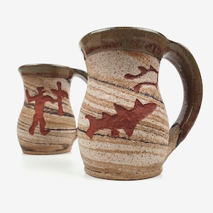 May include: Two ceramic mugs with handles. The mugs feature a beige and brown striped pattern with red tribal-style designs. The rims and handles are a darker brown. The mugs have a rustic, handmade appearance.