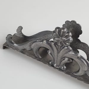 May include: A dark gray metal napkin holder featuring an ornate, raised floral and scrollwork design. The structure has a curved, elongated base with a textured finish and a prominent flower-shaped detail centered on the front panel.