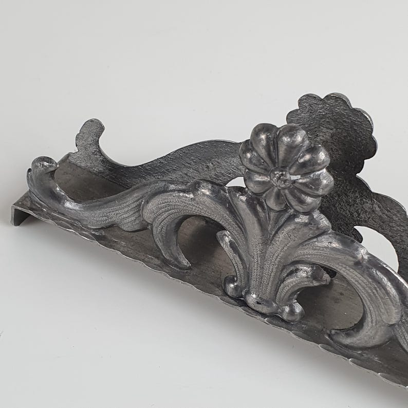 May include: A dark gray metal napkin holder featuring an ornate, raised floral and scrollwork design. The structure has a curved, elongated base with a textured finish and a prominent flower-shaped detail centered on the front panel.
