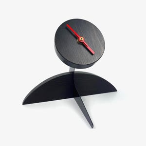 Puede incluir: Un moderno reloj de escritorio negro con esfera circular y manecillas rojas. El reloj tiene un diseño único con una base curva y un soporte delgado. La esfera del reloj tiene aproximadamente 10 cm de diámetro.