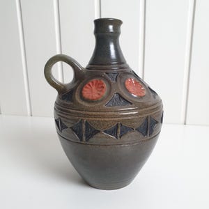 May include: A dark brown ceramic jug with a single handle and a narrow neck. The body features a band of embossed diamond shapes and a row of circular medallions with reddish-orange floral patterns, all finished with a smooth, glossy glaze.