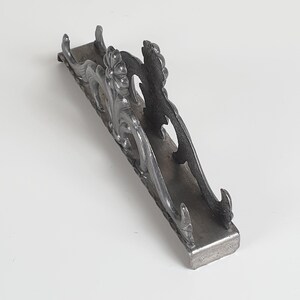 May include: A dark gray metal napkin holder featuring an ornate, raised floral and scrollwork design along the sides. The structure consists of a flat rectangular base with two upright, decorative panels that curve outward at the ends.