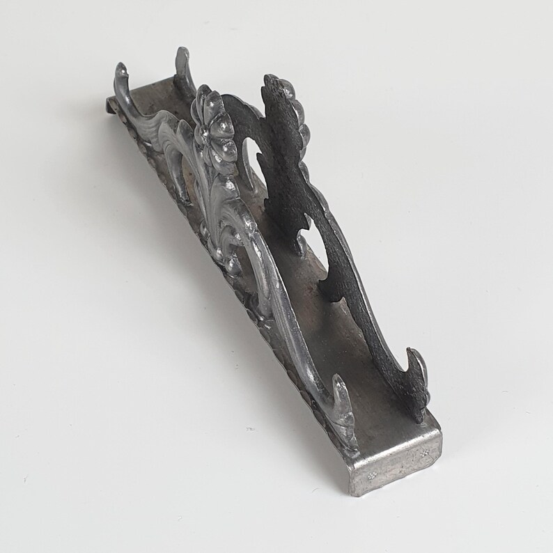 May include: A dark gray metal napkin holder featuring an ornate, raised floral and scrollwork design along the sides. The structure consists of a flat rectangular base with two upright, decorative panels that curve outward at the ends.