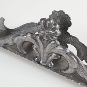 May include: A silver-toned metal napkin holder featuring an ornate, raised floral motif at the center. The structure displays symmetrical, curved scrollwork patterns along the sides with a textured, matte finish throughout the entire piece.