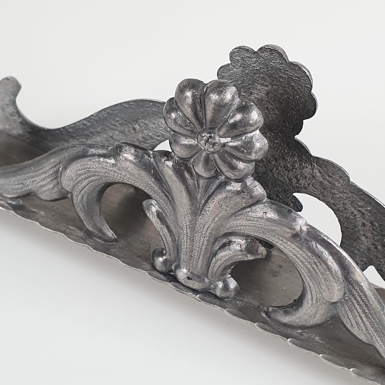 May include: A silver-toned metal napkin holder featuring an ornate, raised floral motif at the center. The structure displays symmetrical, curved scrollwork patterns along the sides with a textured, matte finish throughout the entire piece.