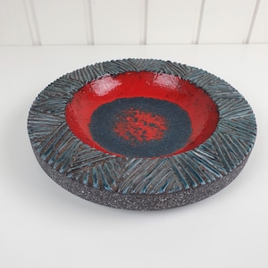 Pode incluir: Uma tigela de cerâmica redonda com um centro vermelho vibrante rodeado por uma base azul escura salpicada. A borda larga e texturizada exibe um padrão geométrico repetitivo de linhas gravadas em tons de azul escuro e cinza com acabamento rugoso.