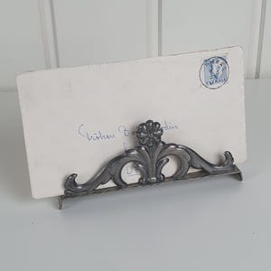 May include: A silver-toned metal stand with an ornate floral and scrollwork design holding a white rectangular envelope. The envelope features blue cursive handwriting and a circular blue postage stamp with the text 25 ORE SVERIGE.