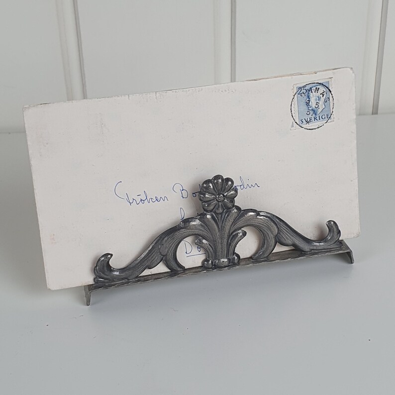 May include: A silver-toned metal stand with an ornate floral and scrollwork design holding a white rectangular envelope. The envelope features blue cursive handwriting and a circular blue postage stamp with the text 25 ORE SVERIGE.