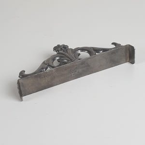May include: A silver-toned metal stand featuring an ornate, raised floral and scrollwork design along the top edge. The base is a flat, rectangular bar with small side tabs. The text "MADE IN JAPAN" and "STV" is stamped on the front surface.