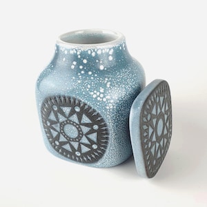 May include: A blue ceramic jar with a matching lid. The jar has a speckled finish and a geometric design in black. The lid is rectangular with a similar design. The jar is approximately 13 cm tall.