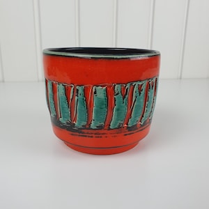 May include: A ceramic planter with a vibrant orange glazed exterior featuring a band of vertical, textured teal green stripes. The interior of the pot is finished in a dark black glaze, and the base has a slightly tapered, stepped design.