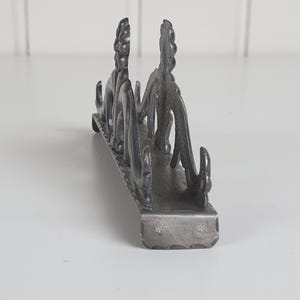 May include: A dark gray metal napkin holder featuring an intricate, raised floral and scrollwork design along its sides. The base is rectangular with a textured edge and small, stamped flower patterns visible on the front-facing end panel.