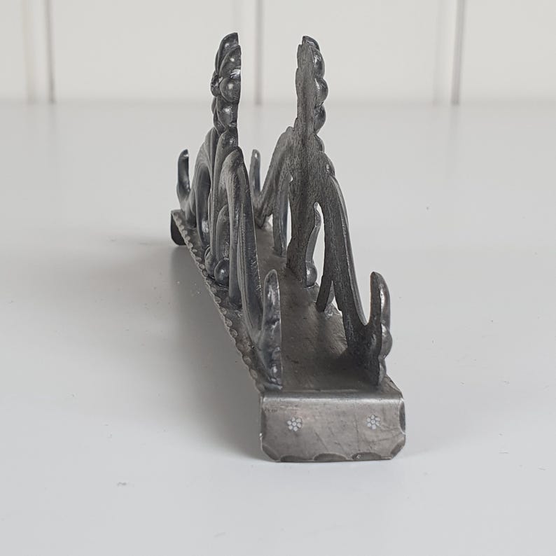 May include: A dark gray metal napkin holder featuring an intricate, raised floral and scrollwork design along its sides. The base is rectangular with a textured edge and small, stamped flower patterns visible on the front-facing end panel.