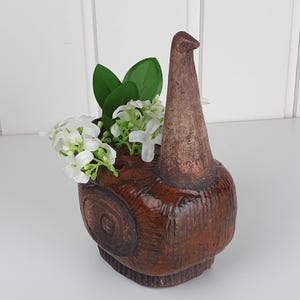 Może przedstawiać: Brązowy ceramiczny wazon w kształcie stylizowanego ślimaka o teksturowanym, ciemnobrązowym wykończeniu. Wazon posiada wysoki, spiczasty występ oraz pusty otwór, w którym znajduje się bukiet sztucznych białych kwiatów z zielonymi liśćmi.