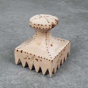 May include: A light-coloured wooden meat tenderiser featuring a square base with sharp, pointed teeth and a rounded handle. The surface is decorated with dark, burned-in dotted lines and small floral patterns, with the letters JRAST visible on top.