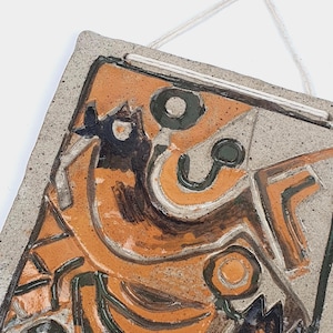 Puede incluir: Un colgante de pared rectangular de cerámica con un diseño abstracto en tonos naranja, negro y verde. La obra de arte presenta formas y líneas geométricas, con una superficie texturizada. Se adjunta una cuerda blanca para colgar.