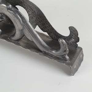 May include: A dark gray, hand-forged metal bracket featuring a textured, hammered surface. The bracket has a curved, scroll-like design with a flat base and a vertical support arm that tapers into a pointed, upward-curving decorative end.