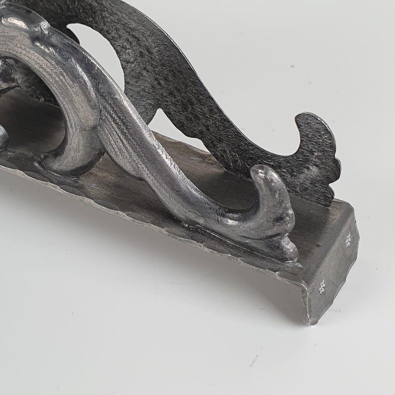May include: A dark gray, hand-forged metal bracket featuring a textured, hammered surface. The bracket has a curved, scroll-like design with a flat base and a vertical support arm that tapers into a pointed, upward-curving decorative end.