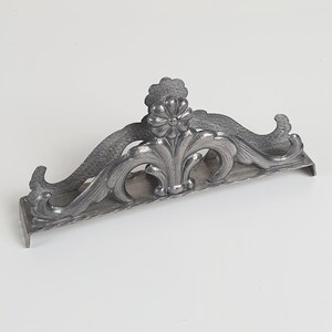 May include: A silver-toned metal napkin holder featuring an ornate, raised floral motif at the center. The design includes symmetrical, curved scrollwork extending outward from the middle flower, with a flat base and two small feet on each end.