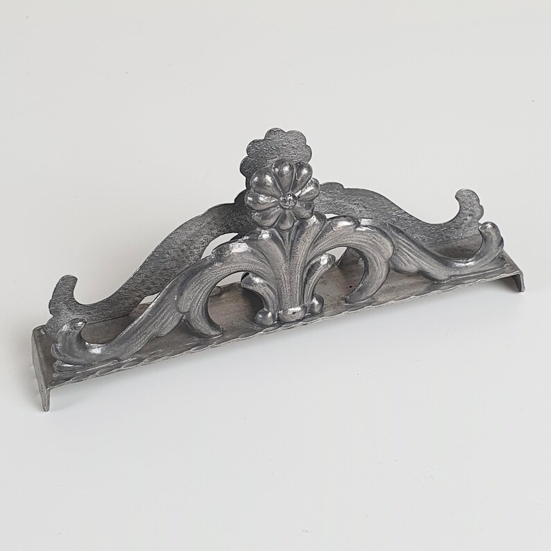 May include: A silver-toned metal napkin holder featuring an ornate, raised floral motif at the center. The design includes symmetrical, curved scrollwork extending outward from the middle flower, with a flat base and two small feet on each end.