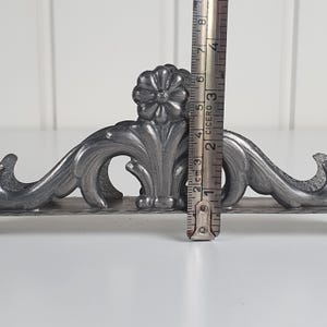 May include: A silver-toned metal stand featuring an ornate floral and scrollwork design. A vertical metal ruler is positioned against the center, showing a height measurement of approximately 8 centimeters for the central decorative element.