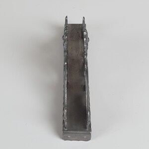 May include: A vertical, dark gray metal napkin holder with a narrow, rectangular base and two upright side panels. The side panels feature a raised, ornate, and textured relief pattern throughout their surface, with a smooth, flat interior spine.
