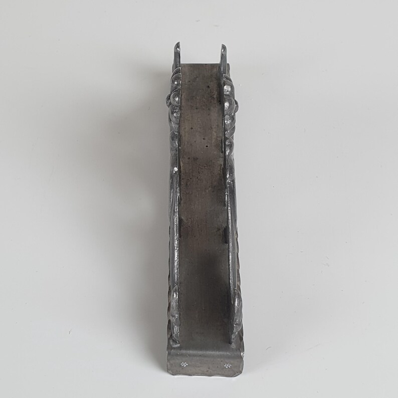 May include: A vertical, dark gray metal napkin holder with a narrow, rectangular base and two upright side panels. The side panels feature a raised, ornate, and textured relief pattern throughout their surface, with a smooth, flat interior spine.