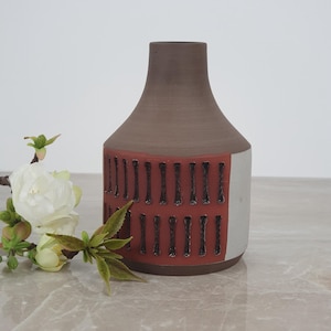 May include: A ceramic vase with a dark brown neck and a wide body featuring a terracotta-colored band decorated with vertical, dark, textured indentations. The lower portion of the vase is finished in a smooth, light gray matte glaze.